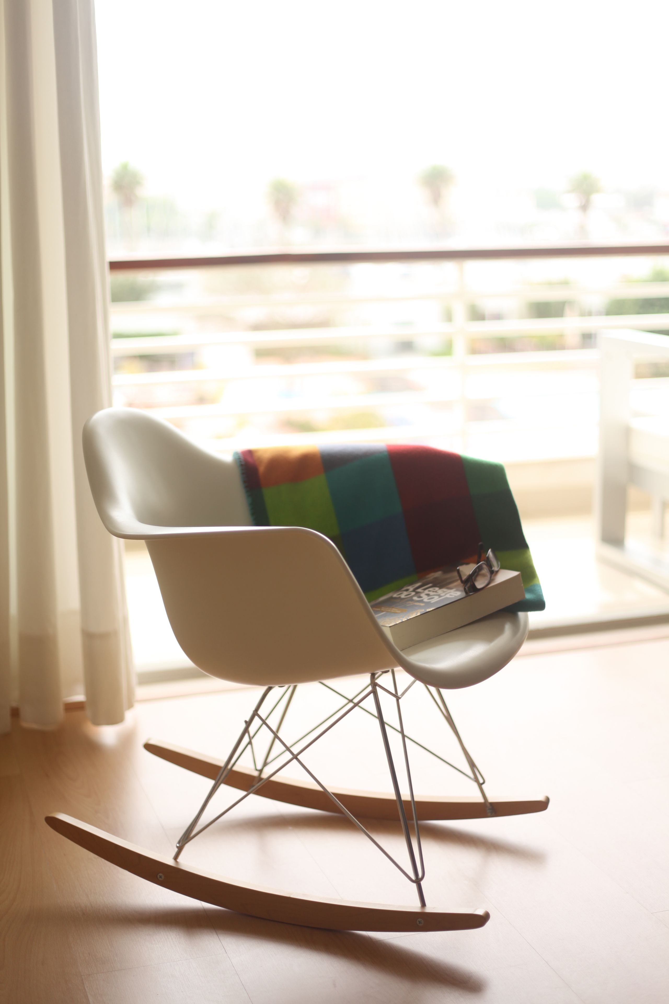 A rocking chair with a colorful blanket, positioned near a window with a view of a balcony.