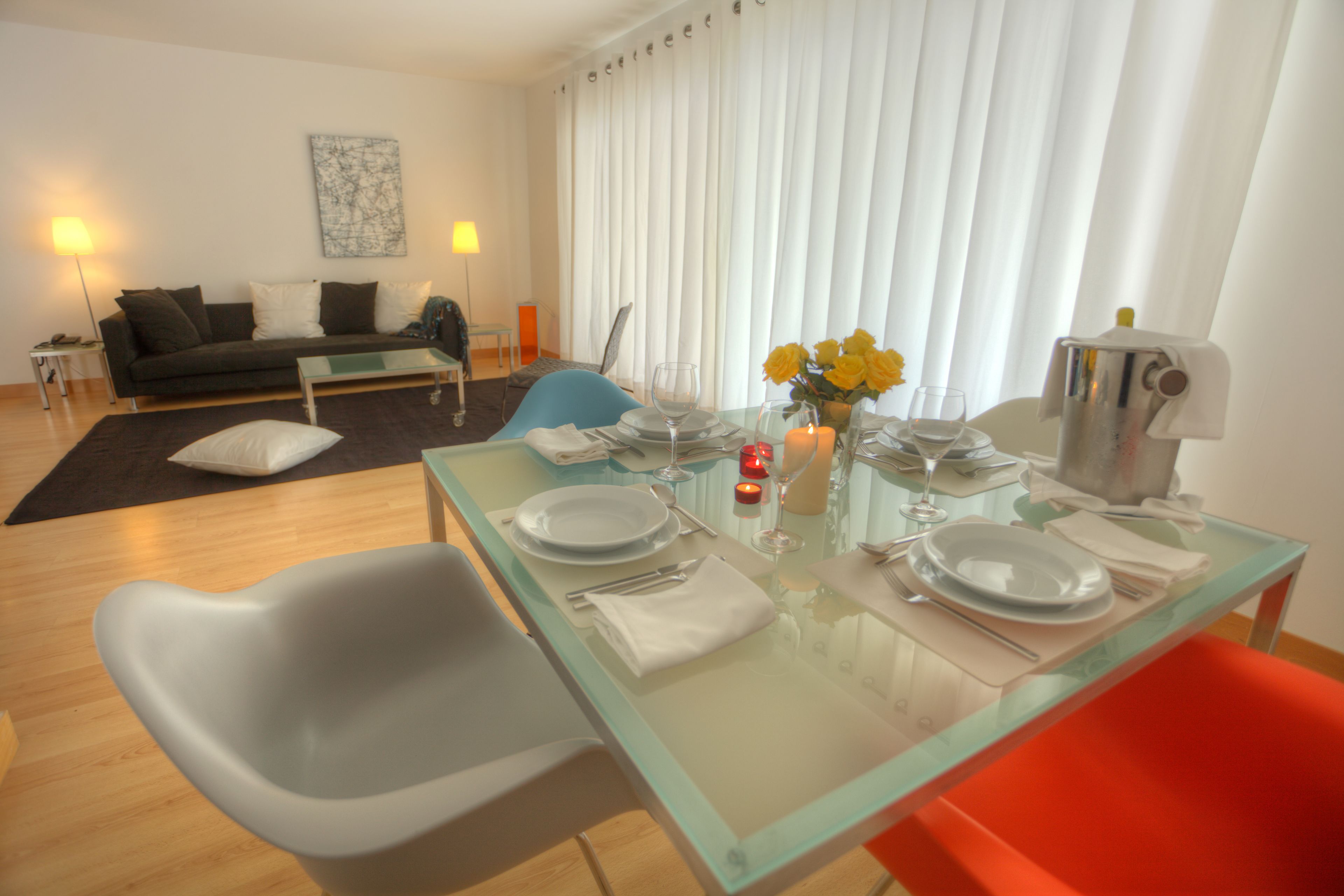 A dining area with a glass table set for four, a sofa, and a lamp in the background.