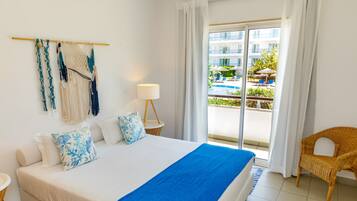 Habitación de hotel con cama, una silla de mimbre, vista a una piscina y edificios, y arte de pared de macramé.