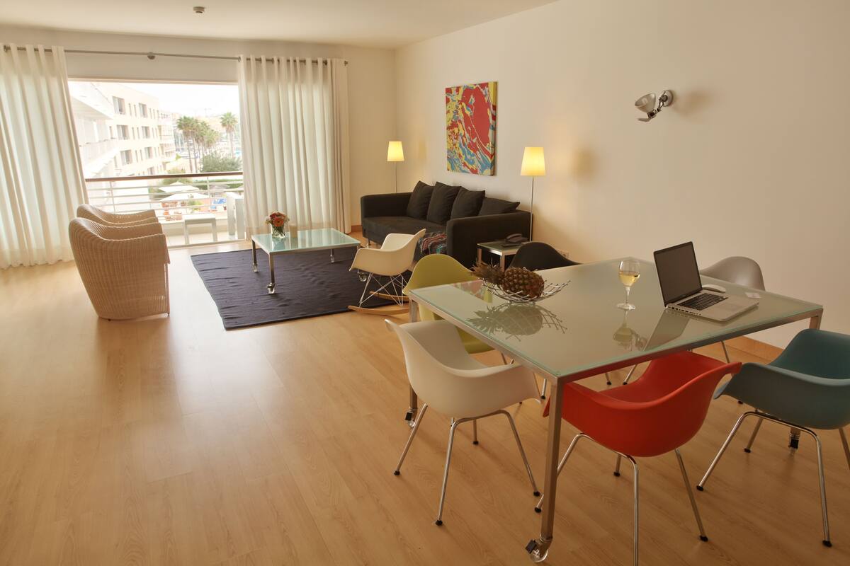 Ein modernes Wohnzimmer mit einem Glastisch, bunten Stühlen, einer Couch und einem Balkon mit Blick auf Gebäude und Palmen.