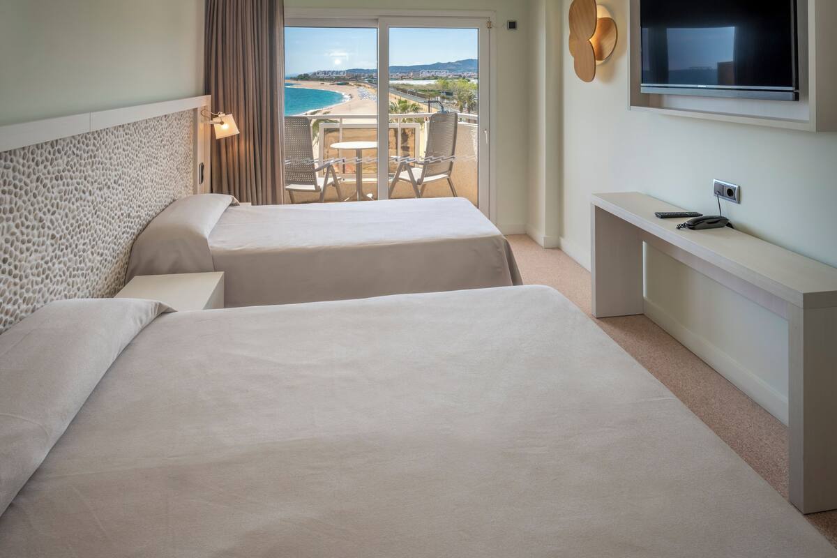 Een hotelkamer met twee bedden, een balkon met een tafel en stoelen, en een aan de muur bevestigde televisie.