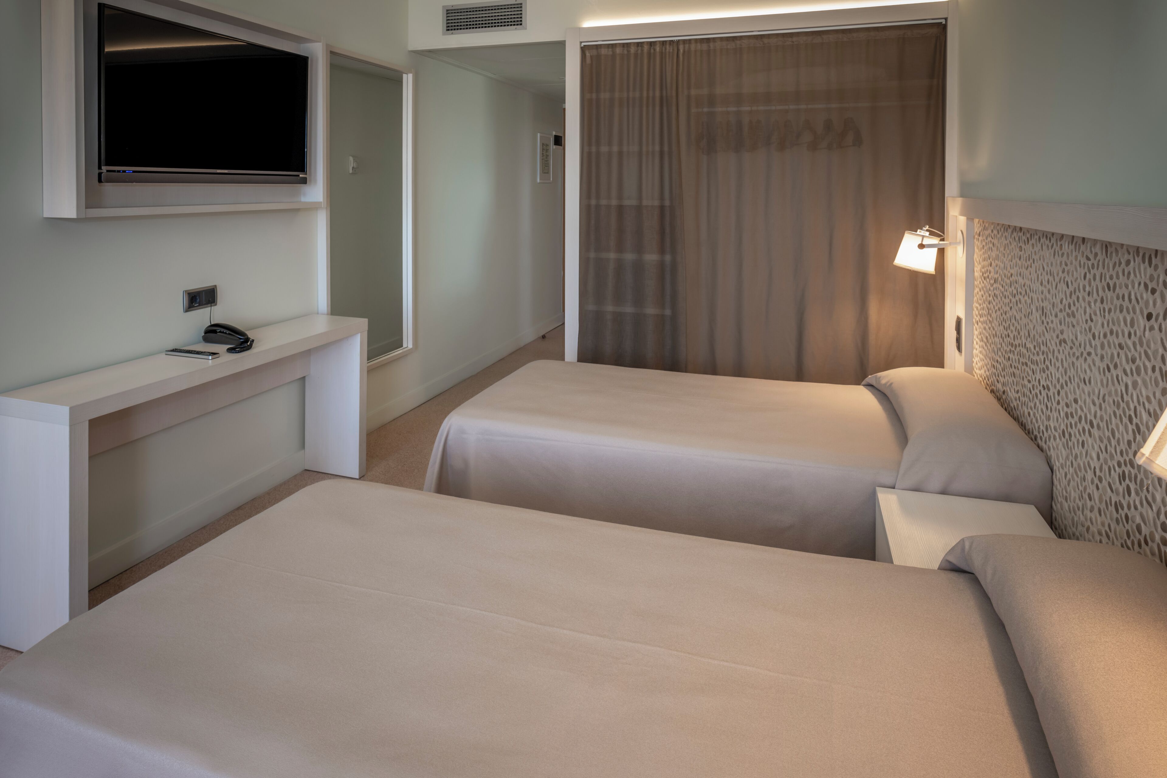 A hotel room with two beds, a TV mounted on the wall, a bedside table with a phone, and a window with curtains.