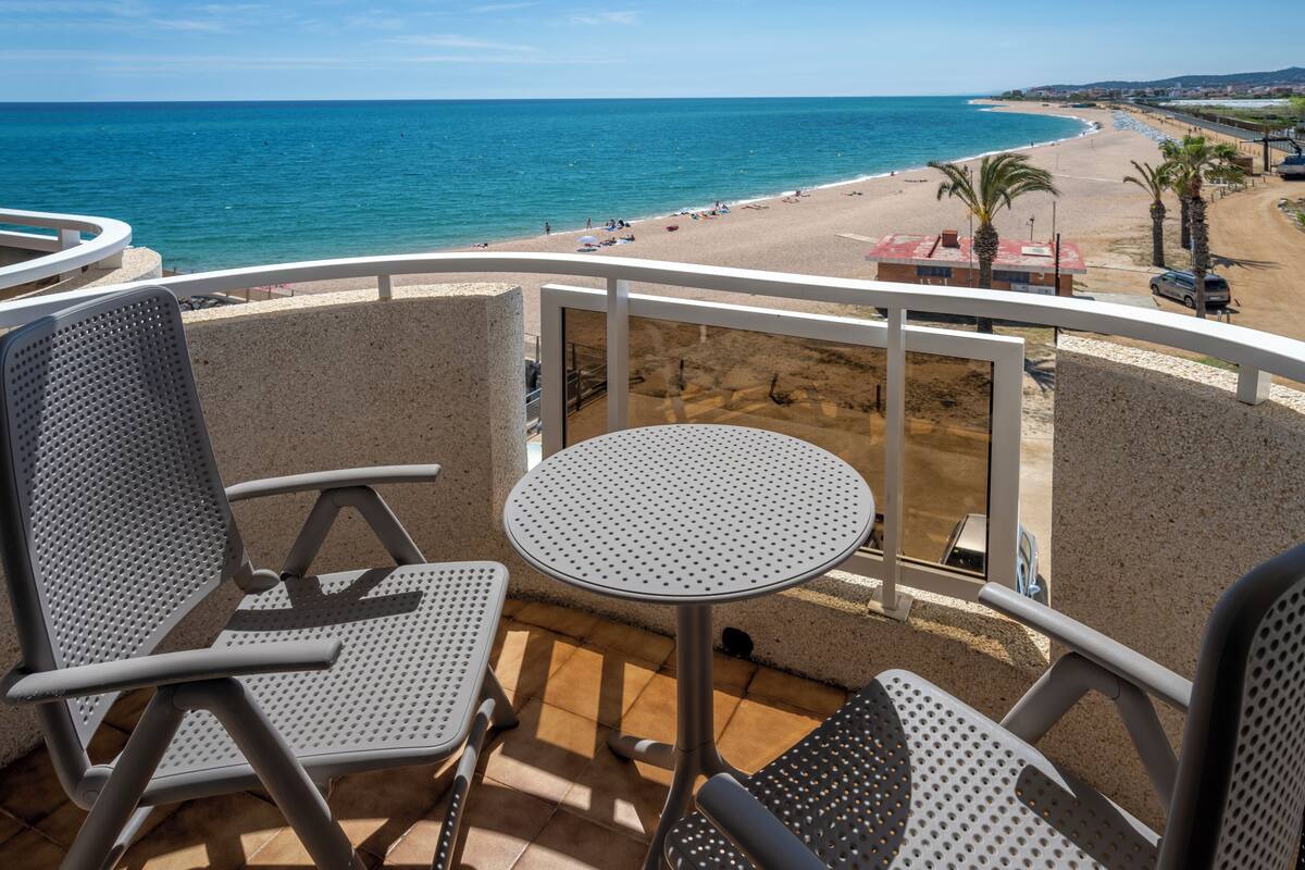 Een balkon met twee stoelen en een tafel, uitkijkend op een strand met palmbomen en een heldere blauwe zee.