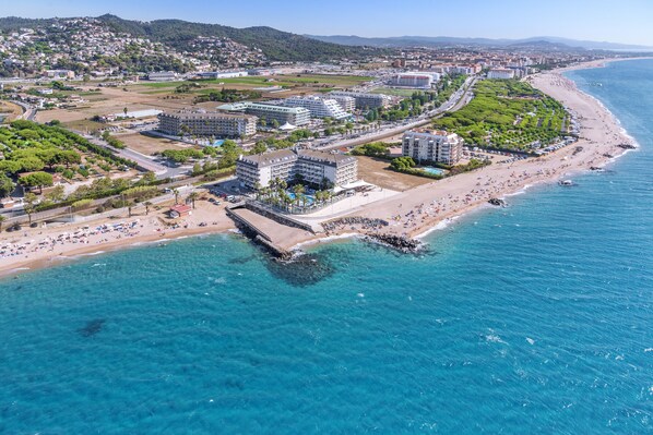 Vista aérea de un hotel costero con varias construcciones, una playa y un paseo marítimo a lo largo del agua.