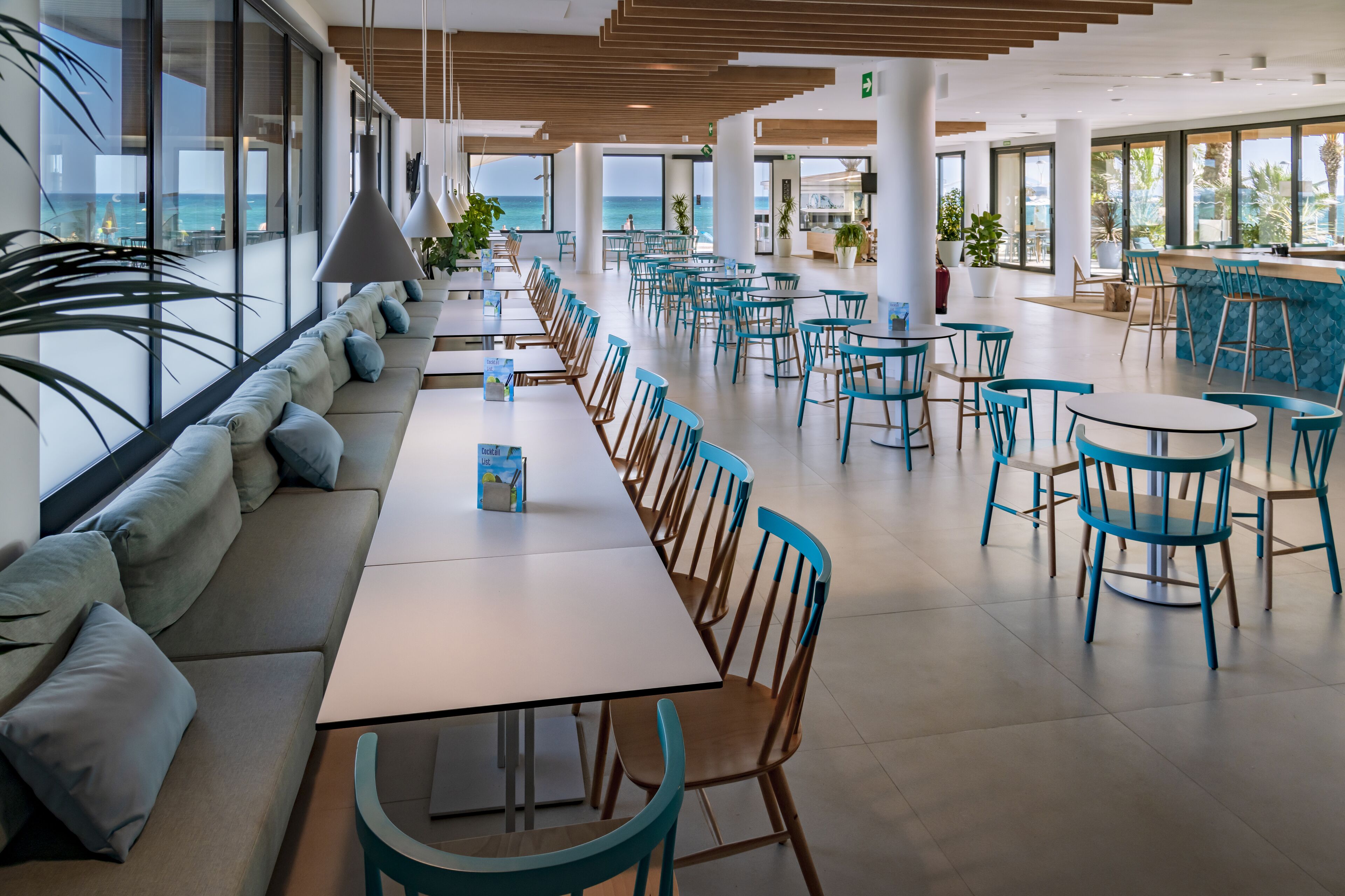 A spacious indoor area with a long table and chairs, large windows with a view of the sea, and potted plants.