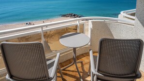 A balcony with two chairs and a small table, overlooking a beach with clear blue water and a rocky shoreline.