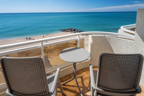 A balcony with two chairs and a small table, overlooking a beach with clear blue water and a rocky shoreline. - Caprici Beach Hotel & Spa (Santa Susanna)