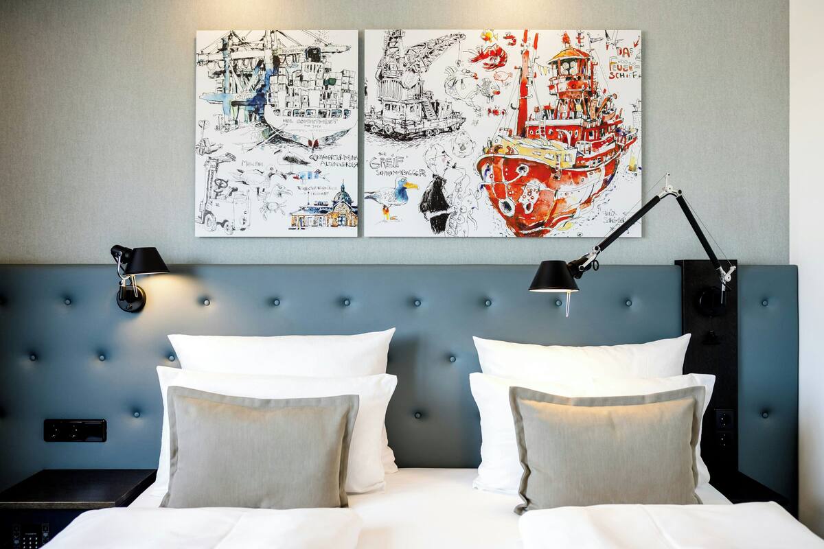 Une chambre d’hôtel avec une tête de lit bleue à motifs capitonnés, du linge de lit blanc et des œuvres d’art représentant des navires et des scènes maritimes accrochées au mur.