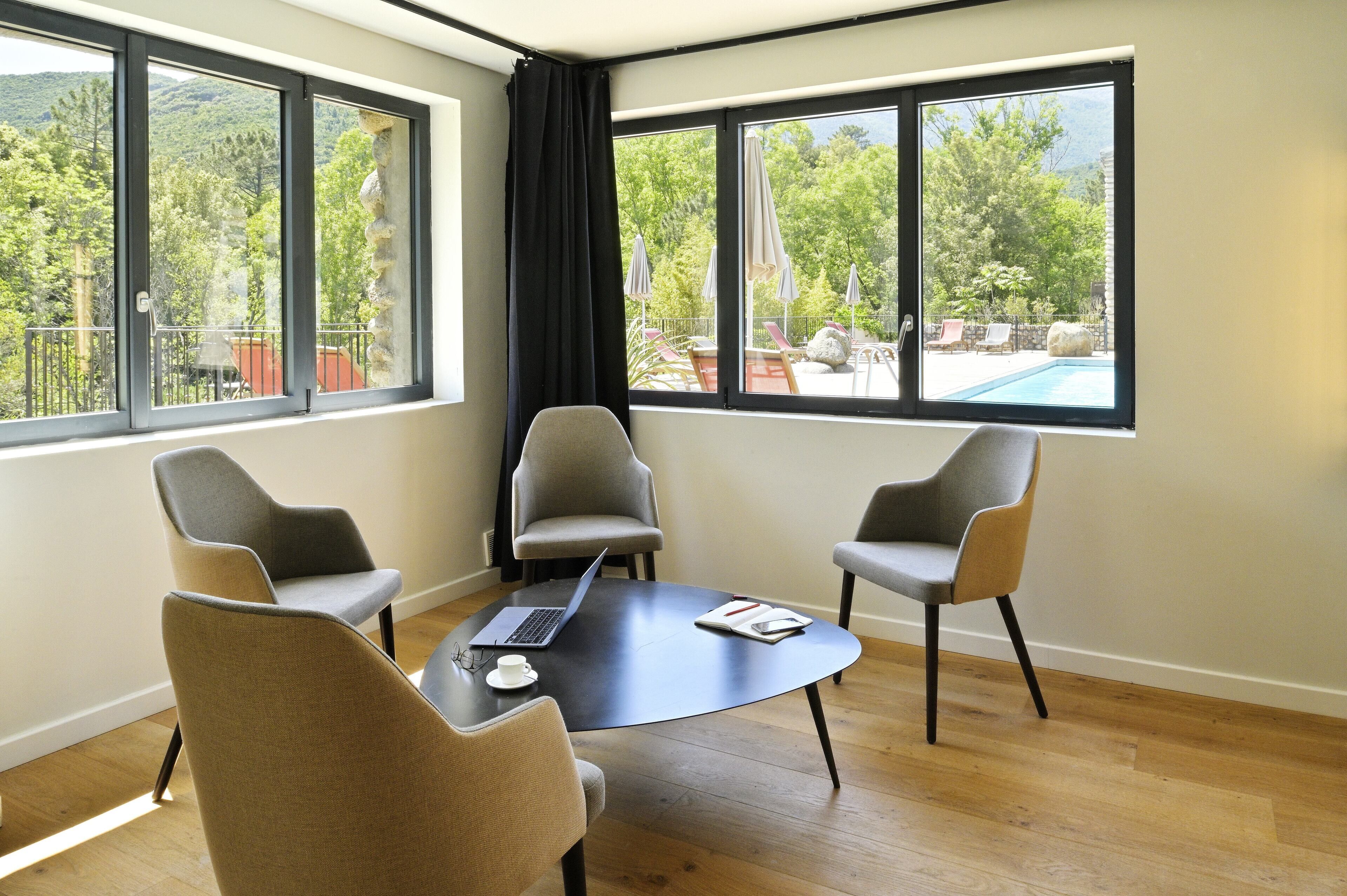 A room with a round table, four chairs, a laptop, and a notebook. Large windows offer a view of a pool and outdoor seating.