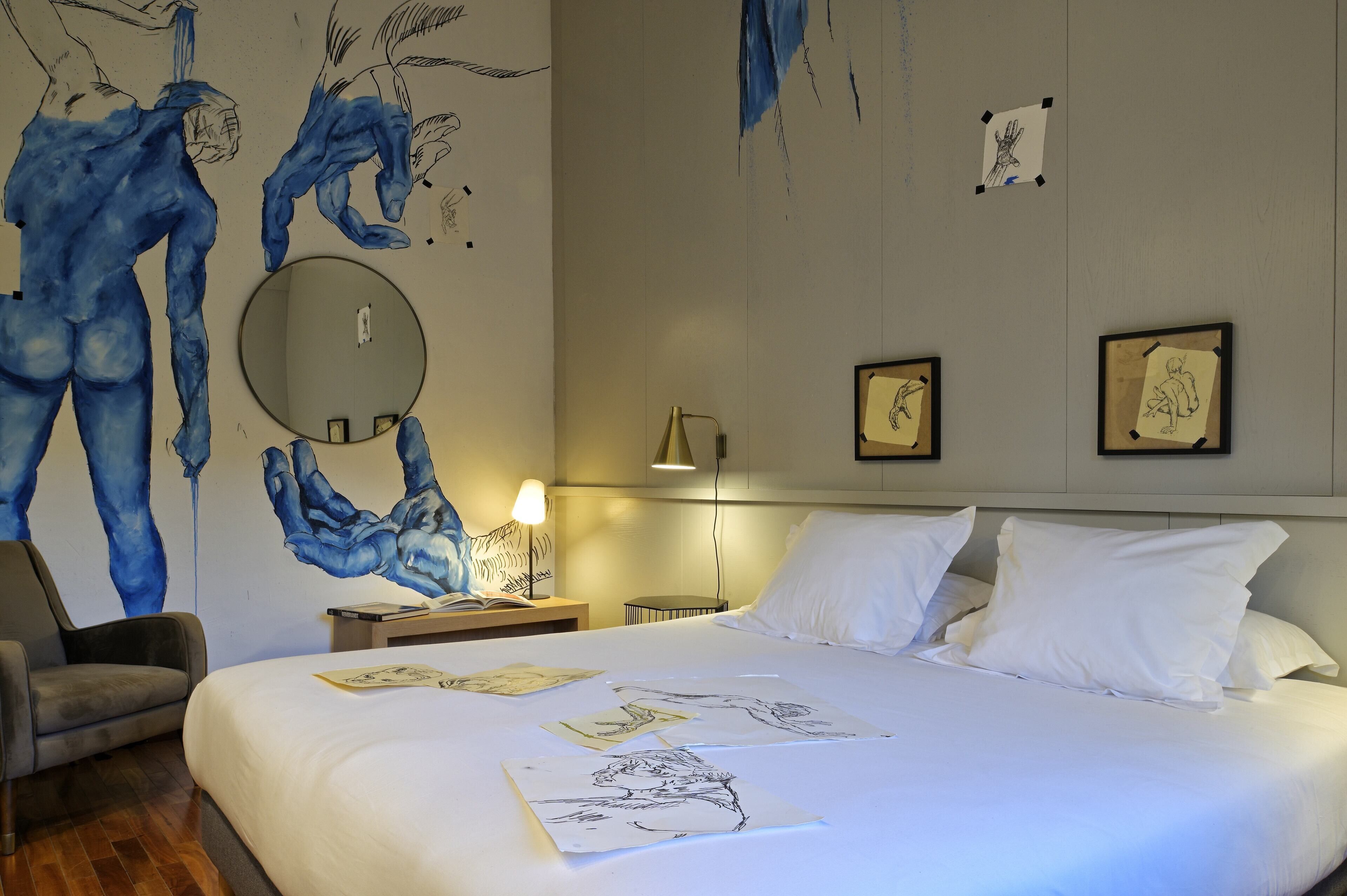 Habitación de hotel con una cama grande, una silla, una mesita de noche con lámpara y una pared decorada con obras de arte.