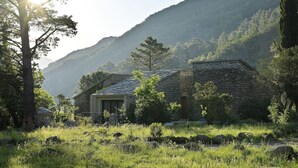 Uma casa de pedra com um pequeno alpendre e uma janela, rodeada por relva e árvores, com montanhas ao fundo.