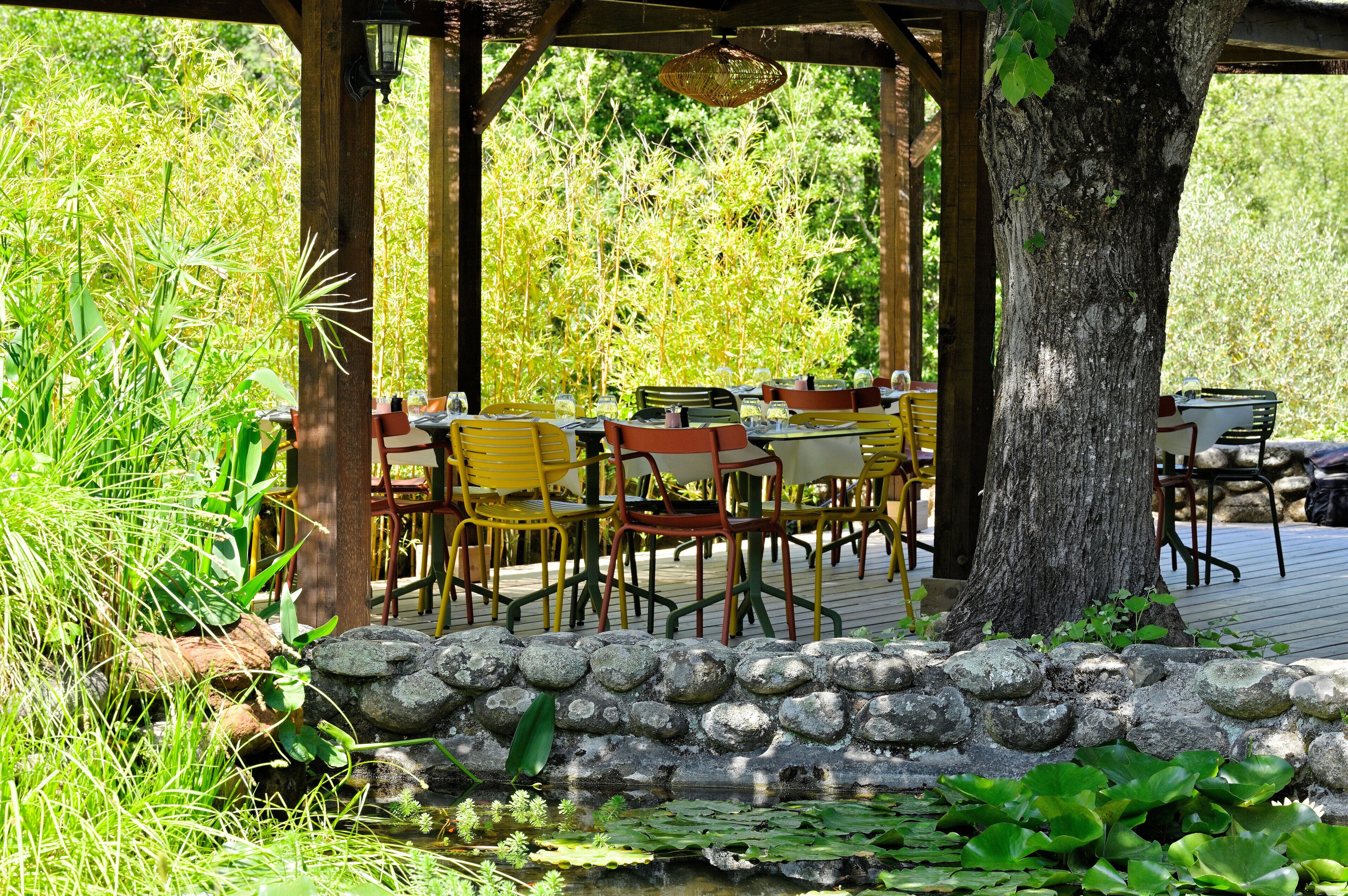 An outdoor dining area with tables and chairs, surrounded by greenery and a stone wall.