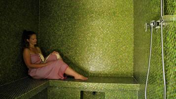 Una mujer sentada en un rincón con bañera de mosaicos, donde se ven una ducha y una manguera.
