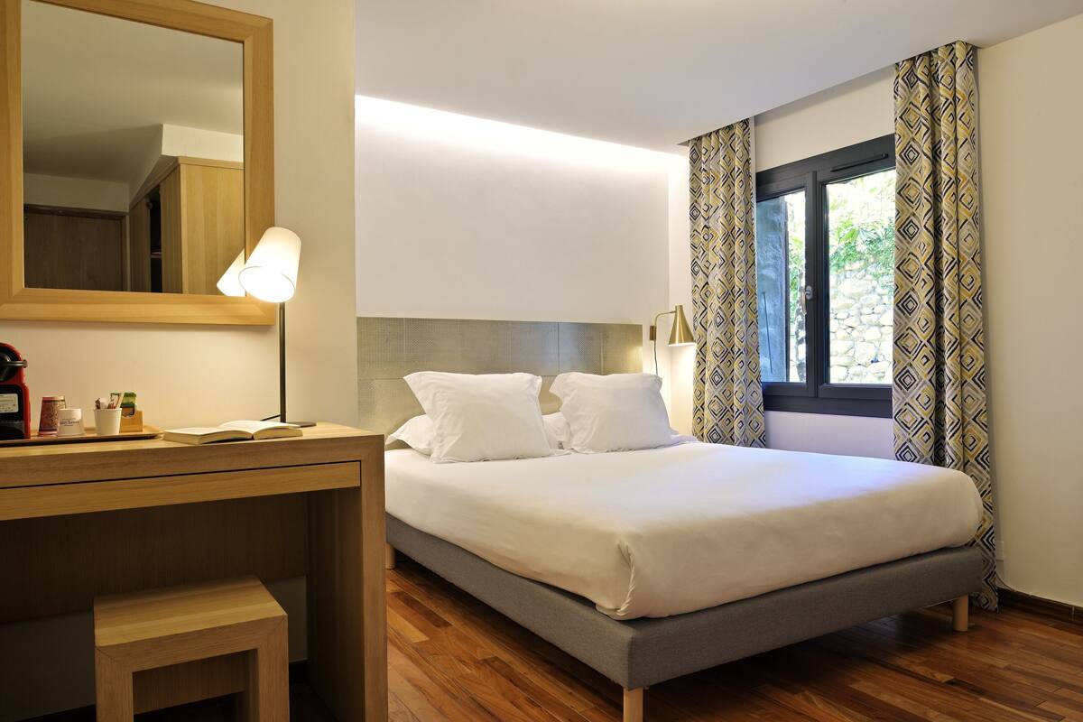 A neatly made bed with white bedding, a wooden desk with a lamp, and a large mirror on the wall.
