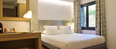Una cama bien tendida con ropa de cama blanca, un escritorio de madera con una lámpara y un espejo grande en la pared.