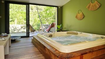 Een spa-ruimte met een bubbelbad, houten terras en uitzicht op een stenen muur en bomen.