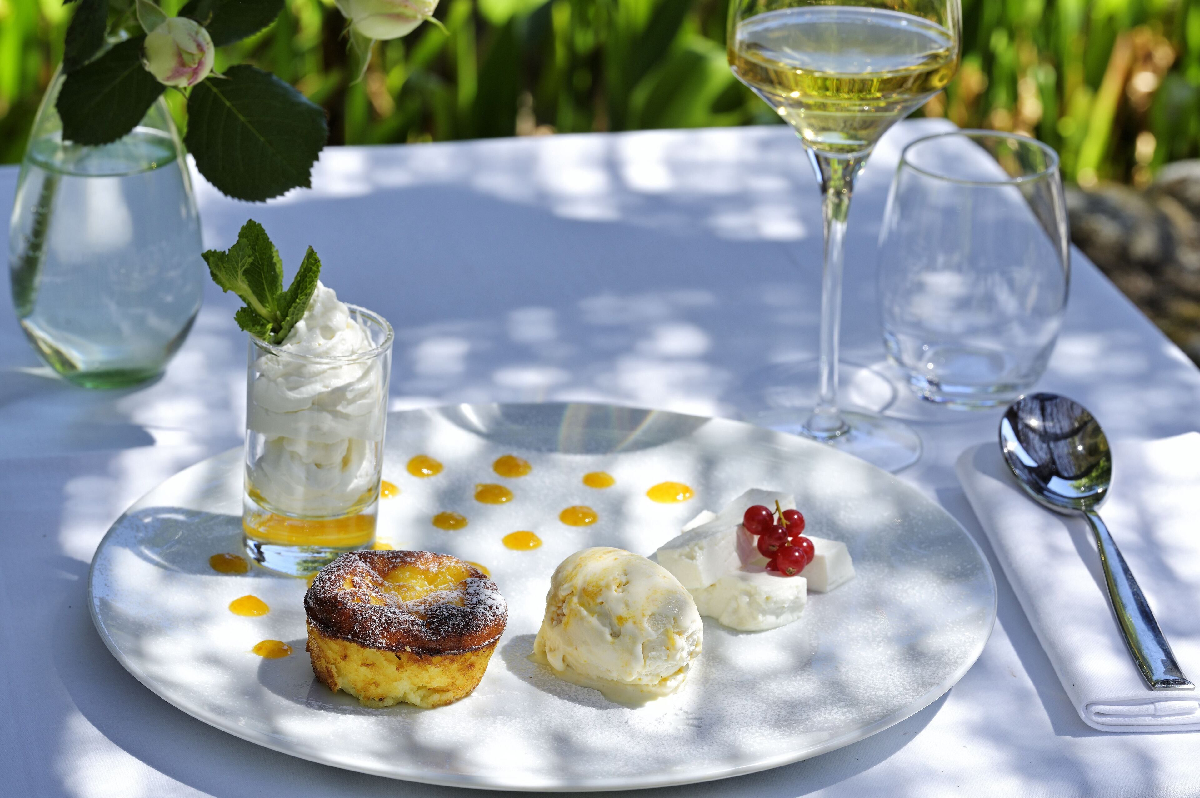 Un plato de postres con varios dulces y una copa de vino blanco sobre una mesa al aire libre.