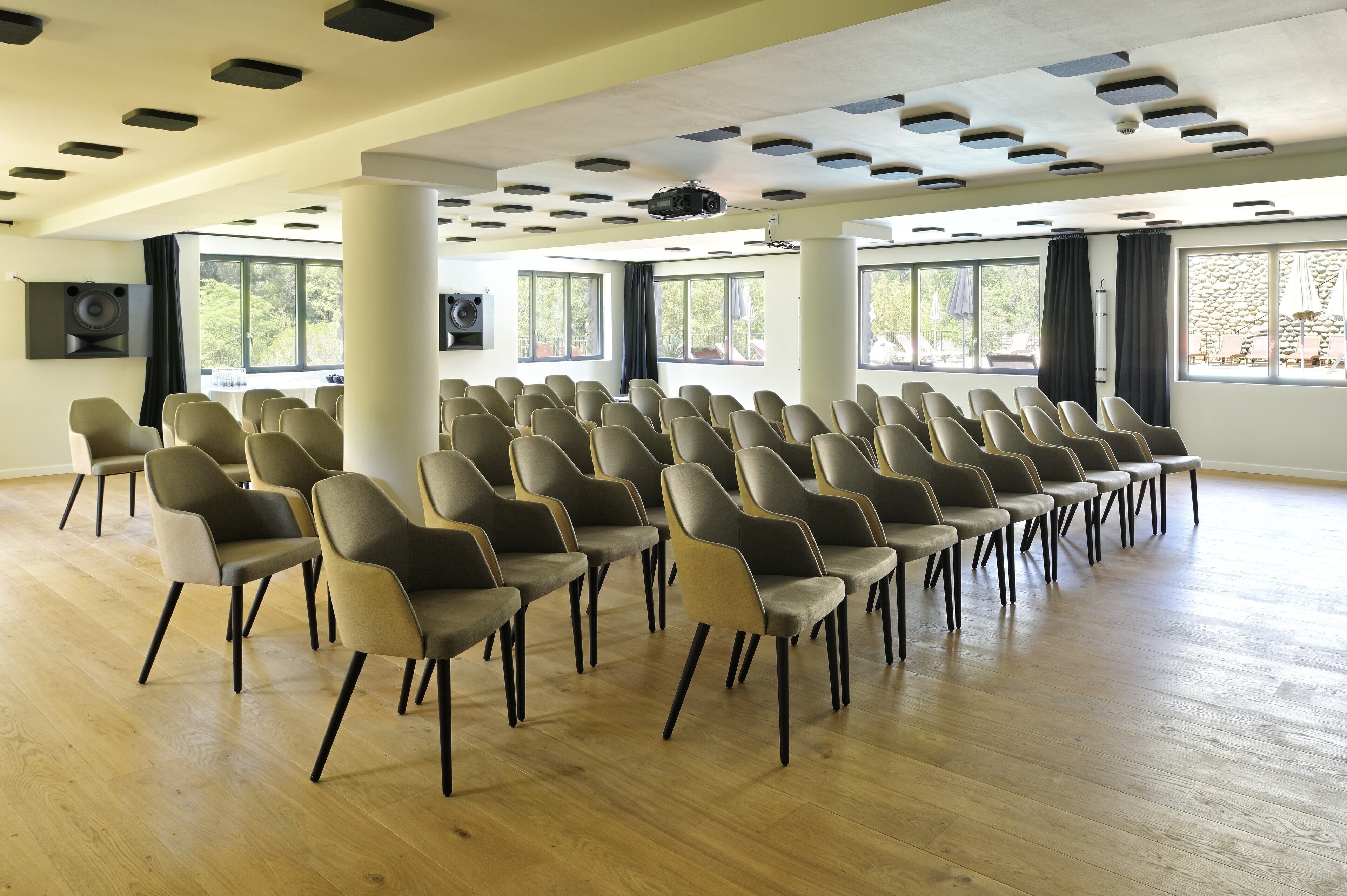 Een vergaderzaal met rijen stoelen, grote ramen, een beamer en een audiosysteem.