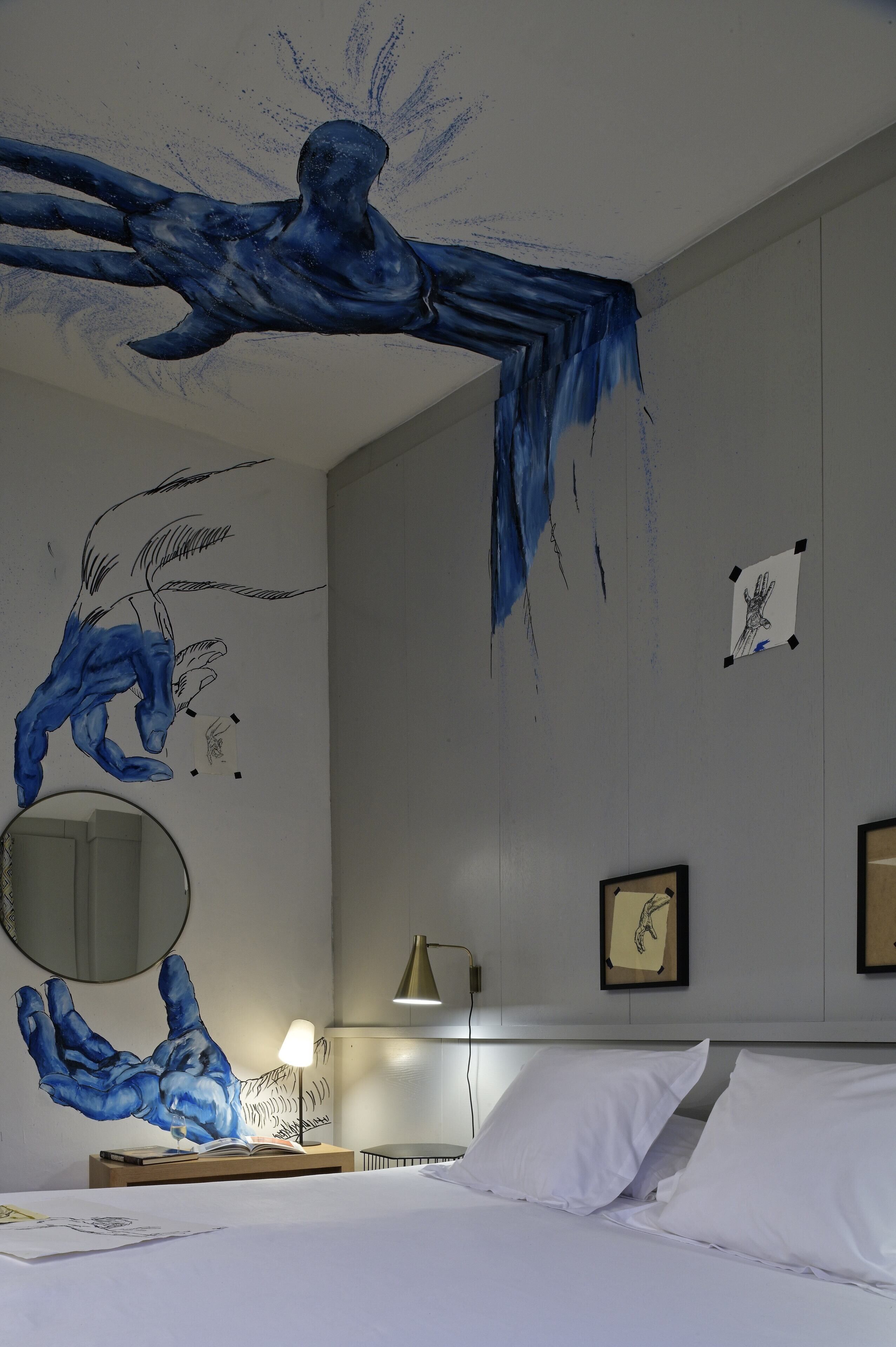 Une chambre d’hôtel avec un lit, une table de chevet avec une lampe, un mur orné d’une grande fresque représentant une empreinte de main bleue, et un tableau encadré accroché au mur.