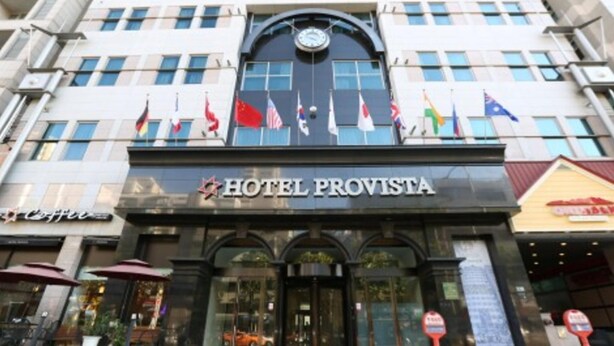 The exterior of Hotel Provista with a clock above the entrance, multiple flags, and a sign indicating a coffee shop nearby. - Provista Hotel (Seoul)