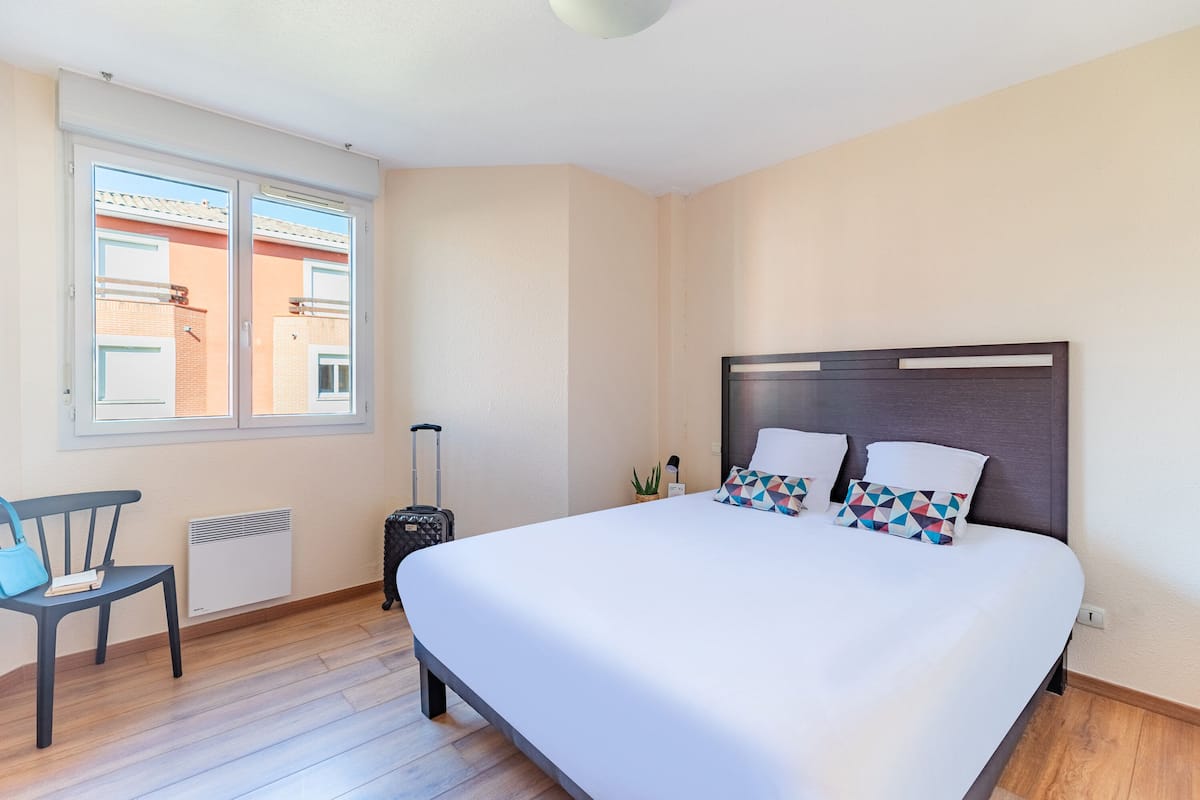 Ett hotellrum med en stor säng, en stol, en resväska och ett fönster med utsikt över byggnaderna utanför.