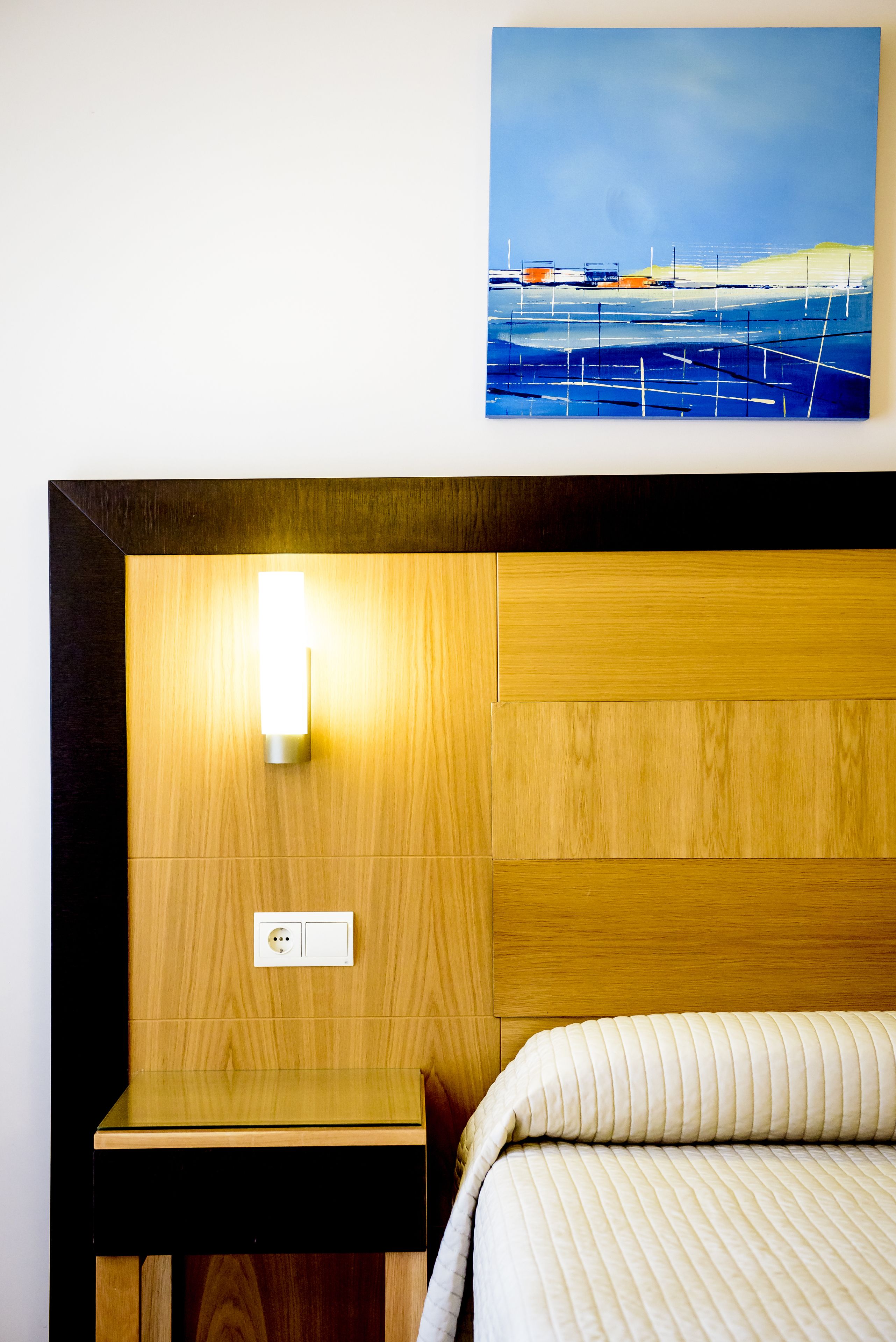 Habitación de hotel con cabecera de madera, una mesita de noche, cama con ropa de cama a rayas y un cuadro con cielo azul y barcos en la pared.