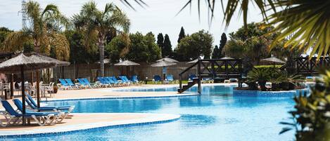 Una piscina con sillas de descanso y sombrillas, rodeada de palmeras y una terraza de madera.