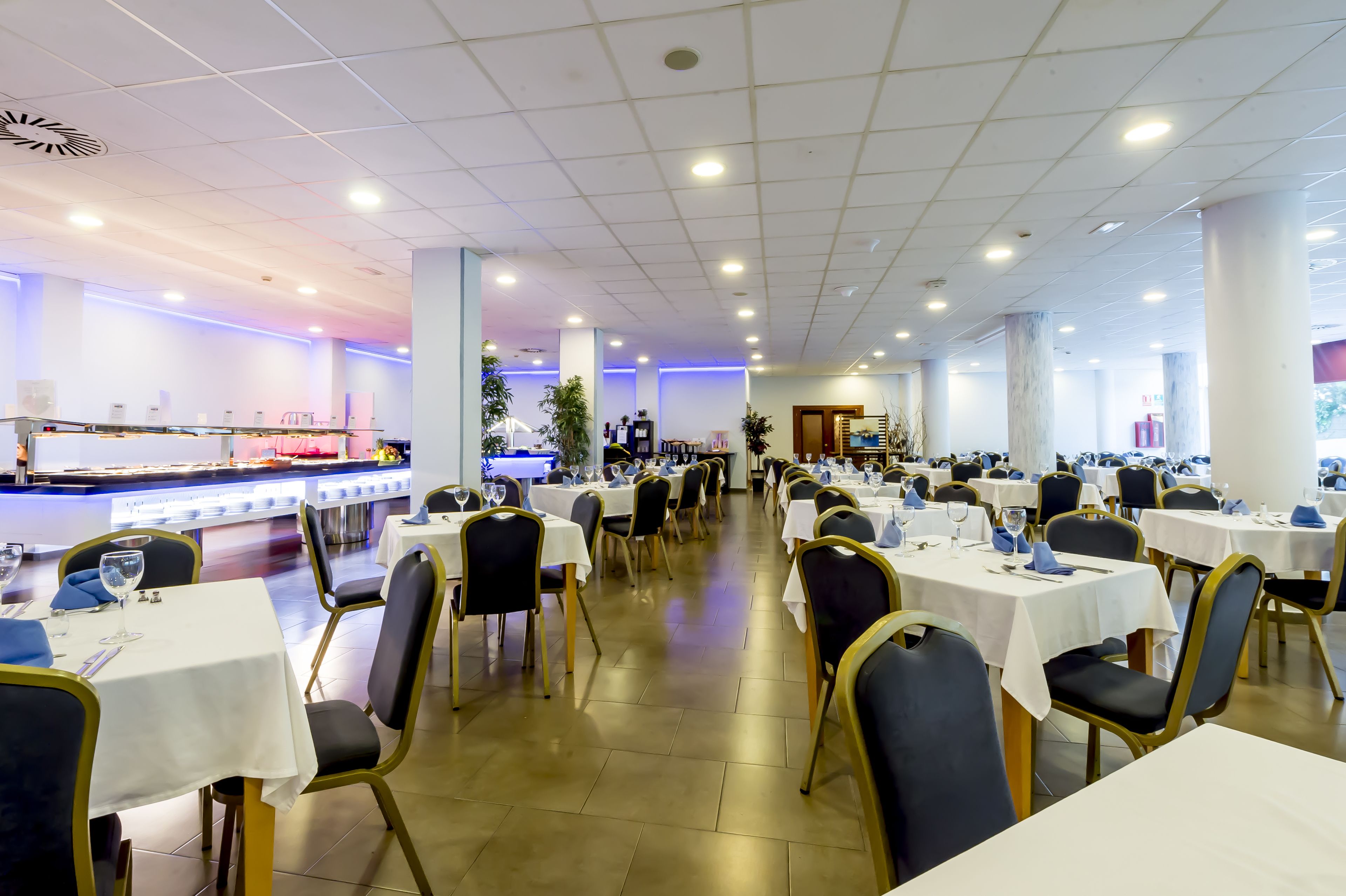 Een ruime eetruimte met meerdere tafels en stoelen, een buffet en een goed verlicht plafond met inbouwspots.