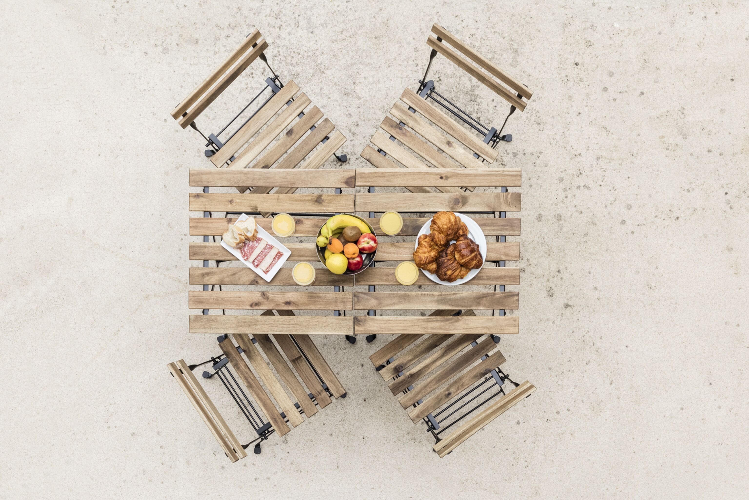 Une table en bois, sur laquelle sont disposés un panier de fruits, des croissants et une assiette de nourriture, le tout posé sur une surface sablonneuse.