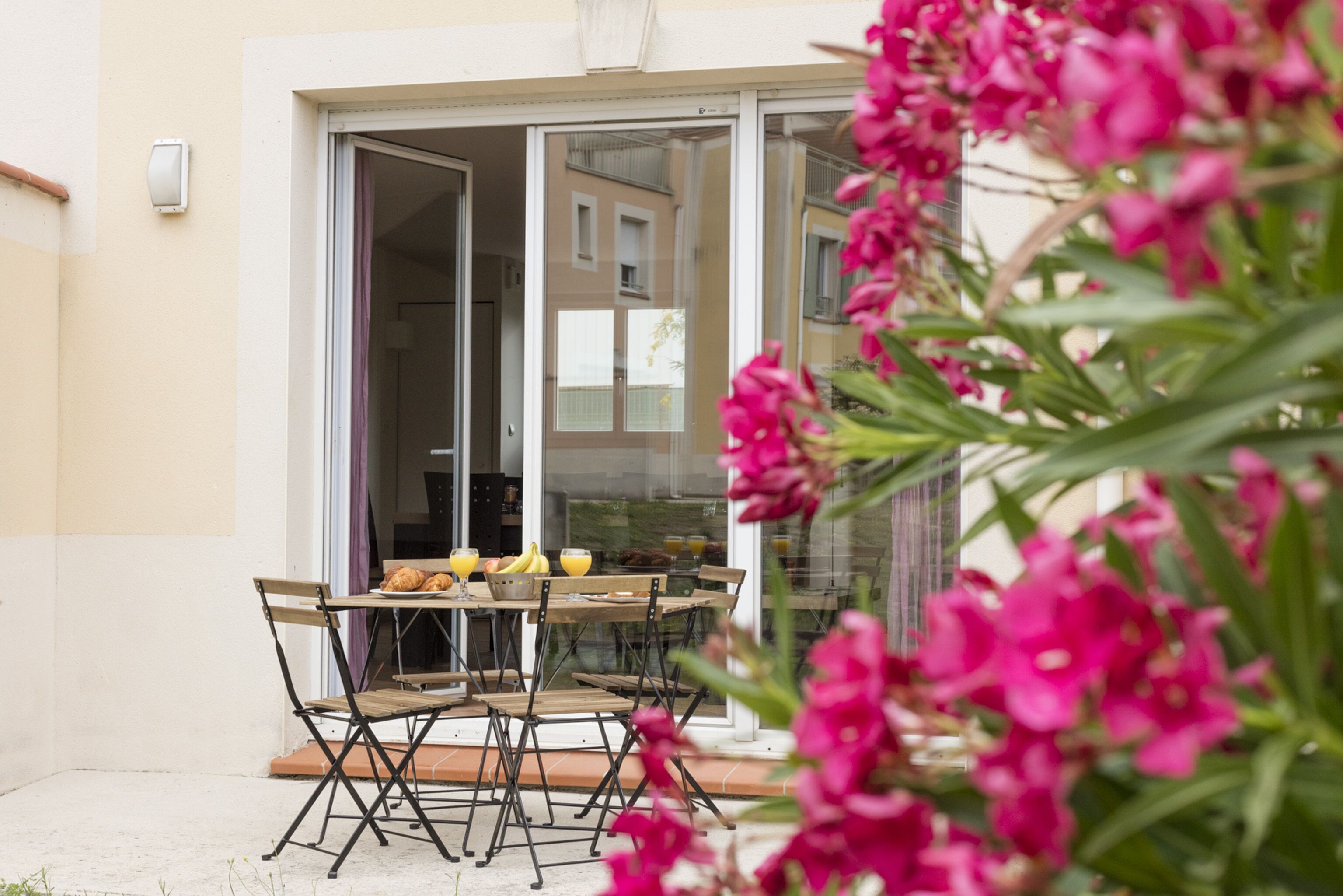 Un espace extérieur aménagé pour les repas, avec une table et des chaises, une porte vitrée donnant sur un balcon, et des fleurs roses au premier plan.