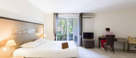 A hotel room with a bed, a desk, a chair, a TV, and a balcony with a view of trees.