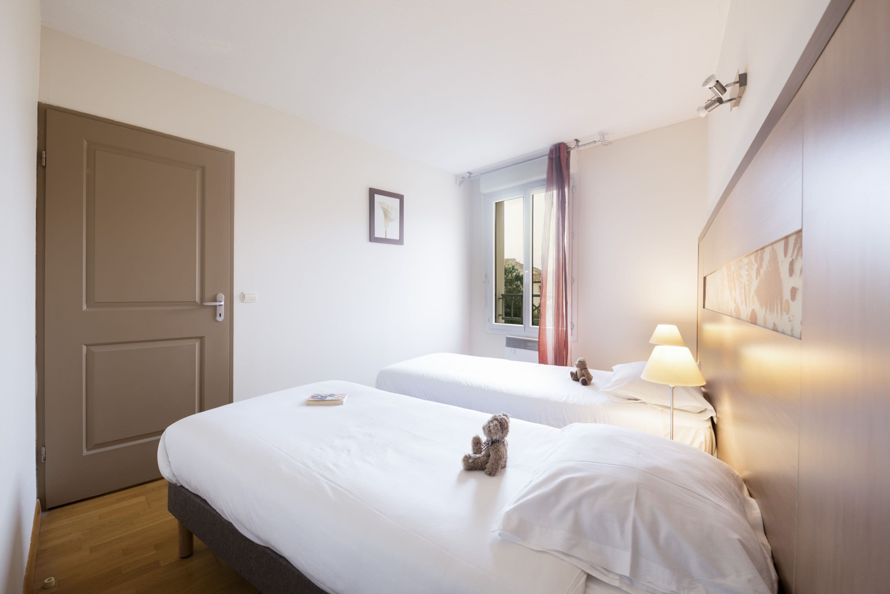 A hotel room with two beds, a door, a window with curtains, a bedside lamp, and a framed picture on the wall.