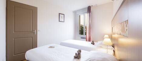 Habitación de hotel con dos camas, una puerta, una ventana con cortinas, una lámpara de noche y un cuadro en la pared.
