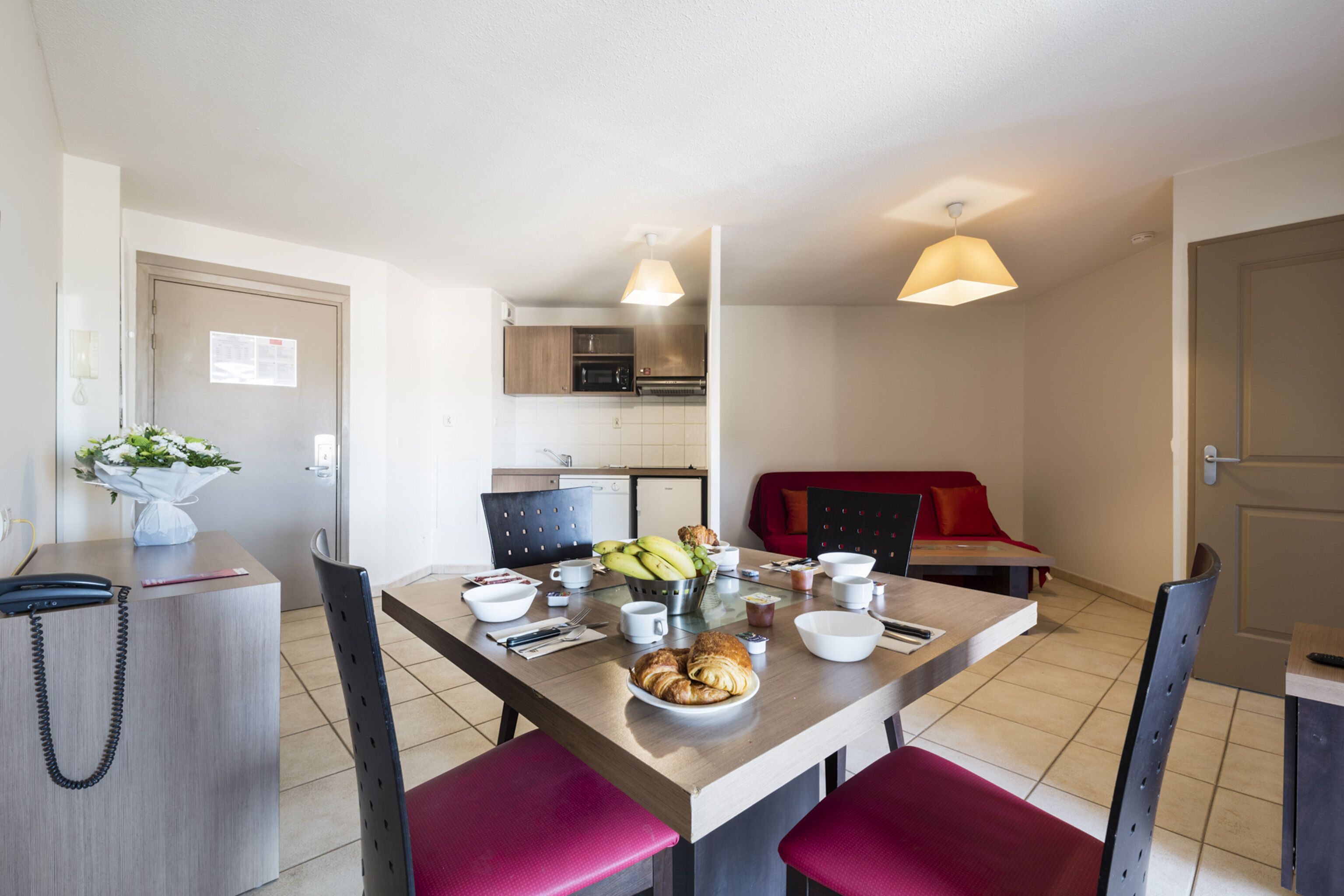 Cocina moderna con zona de comedor, que incluye una mesa servida con comida y tazas, un sofá rojo y un teléfono sobre un escritorio.