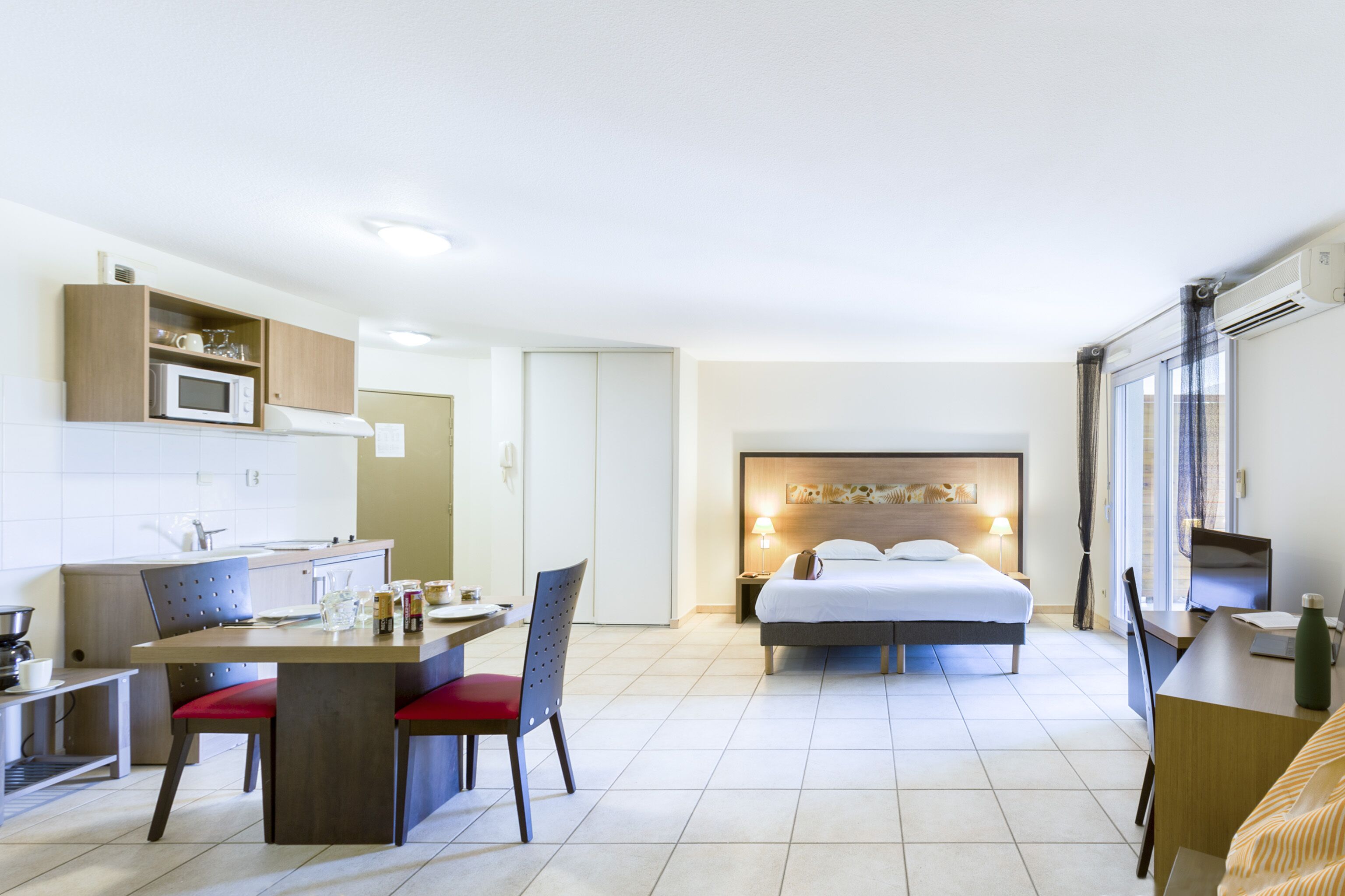 Quarto de hotel com cozinha compacta, cama com cabeceira, escrivaninha com cadeira e uma janela grande.