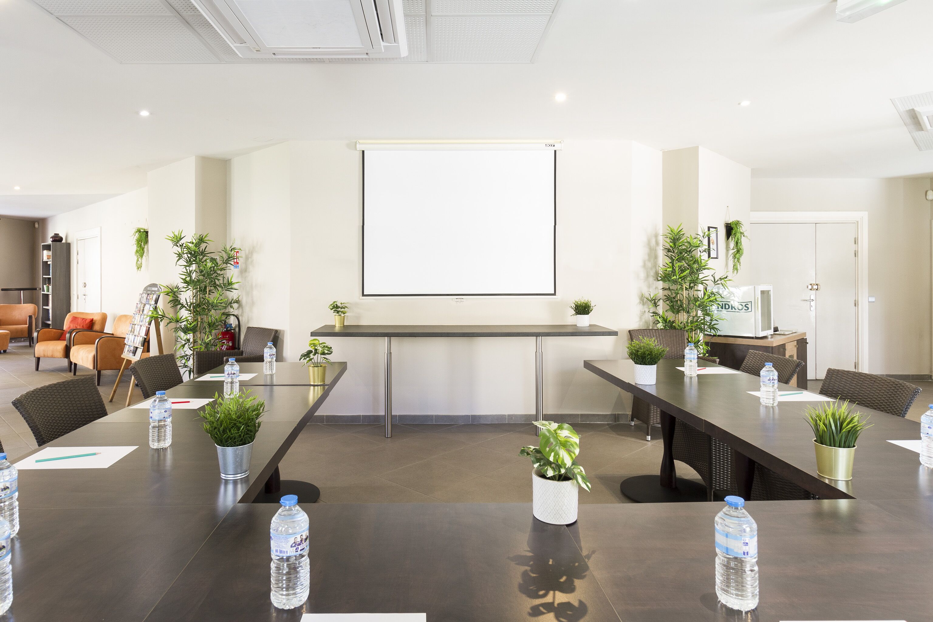 Un salón de conferencias con una larga mesa, sillas, botellas de agua y plantas en macetas.