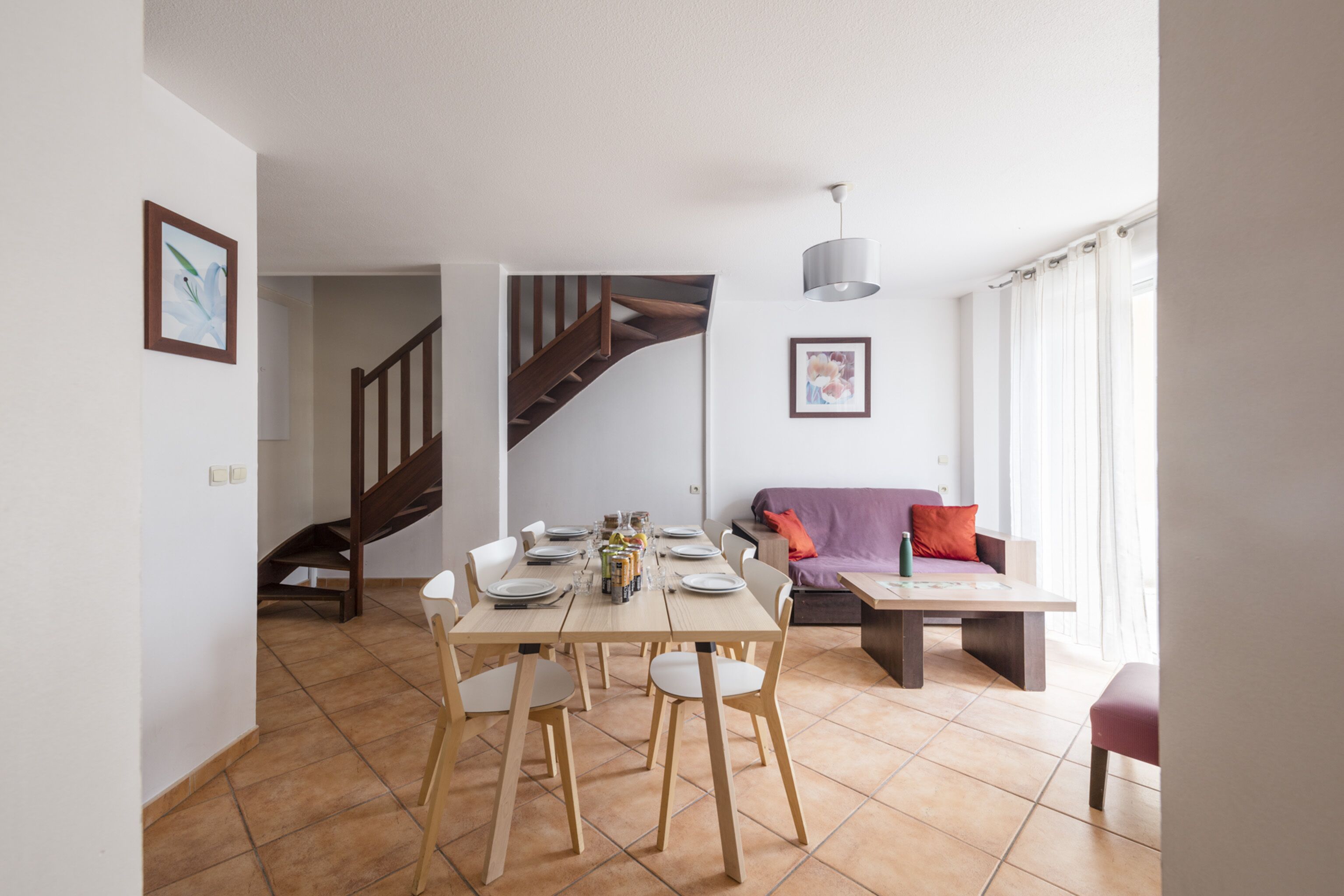 A dining area with a wooden table and chairs, a staircase, a sofa, and a window with curtains.