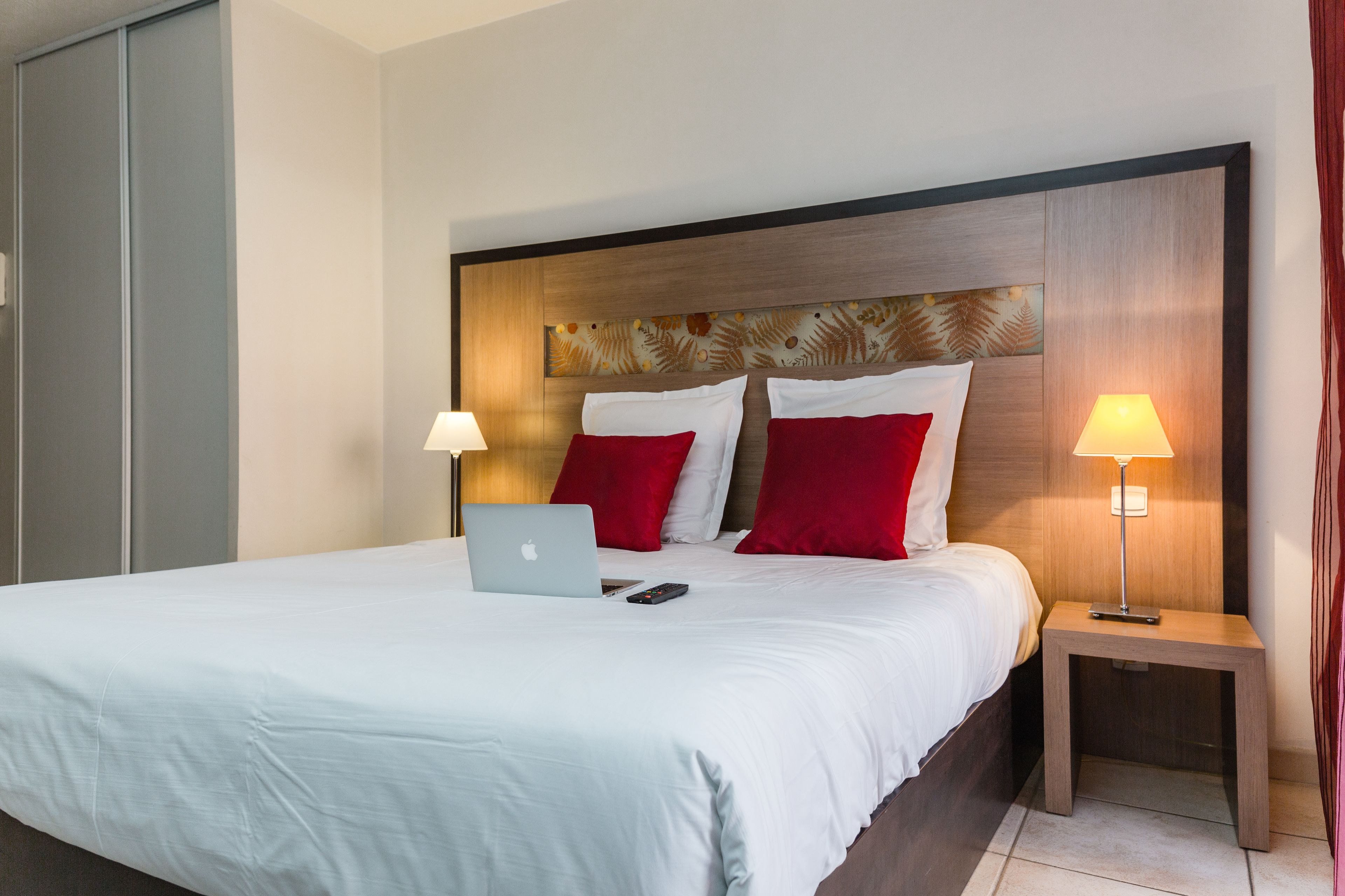 A neatly made bed with a laptop and remote control, a wooden headboard with a leaf pattern, and two bedside lamps.