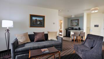 A modern hotel room with a grey sofa, a matching armchair, a coffee table, a floor lamp, and a framed picture on the wall.