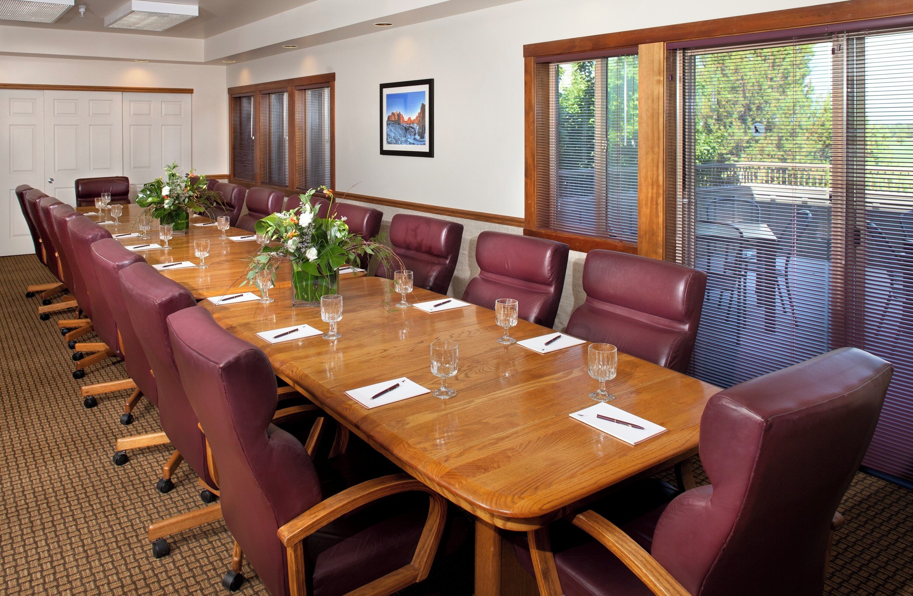 A conference room with a long wooden table, red leather chairs, and a large window with blinds.