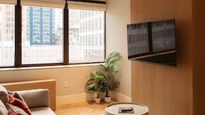 Un salon moderne avec un canapé, une table basse ronde, un téléviseur fixé au mur et une grande fenêtre donnant sur la ville.