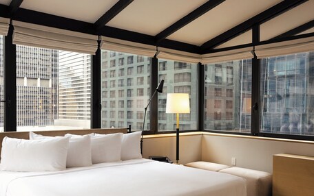 A modern hotel room with a large bed, a bedside table, a lamp, and a view of the city through large windows.