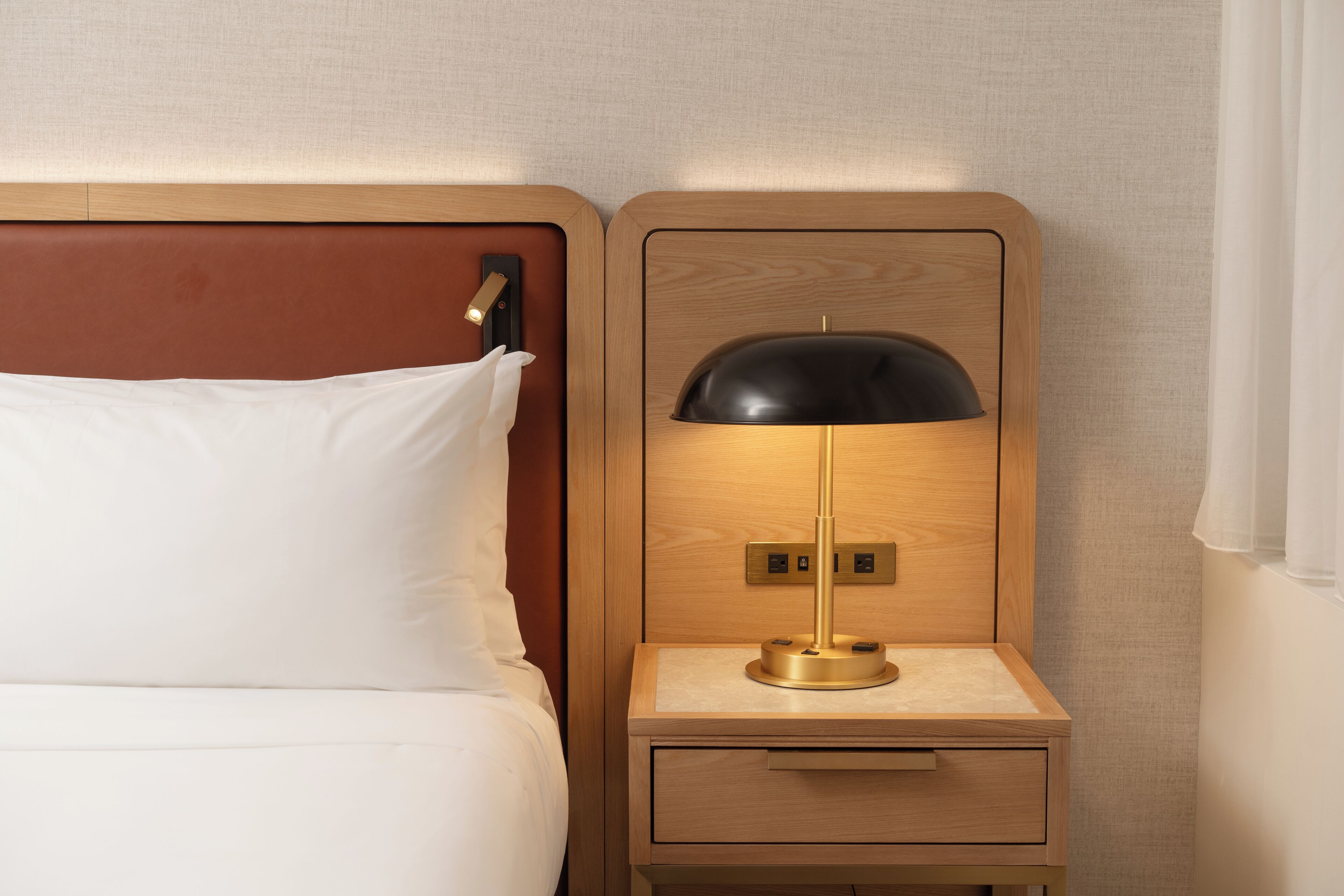 Un lit bien fait, avec du linge de lit blanc, une table de chevet en bois sur laquelle se trouve une lampe, et un interrupteur fixé au mur.