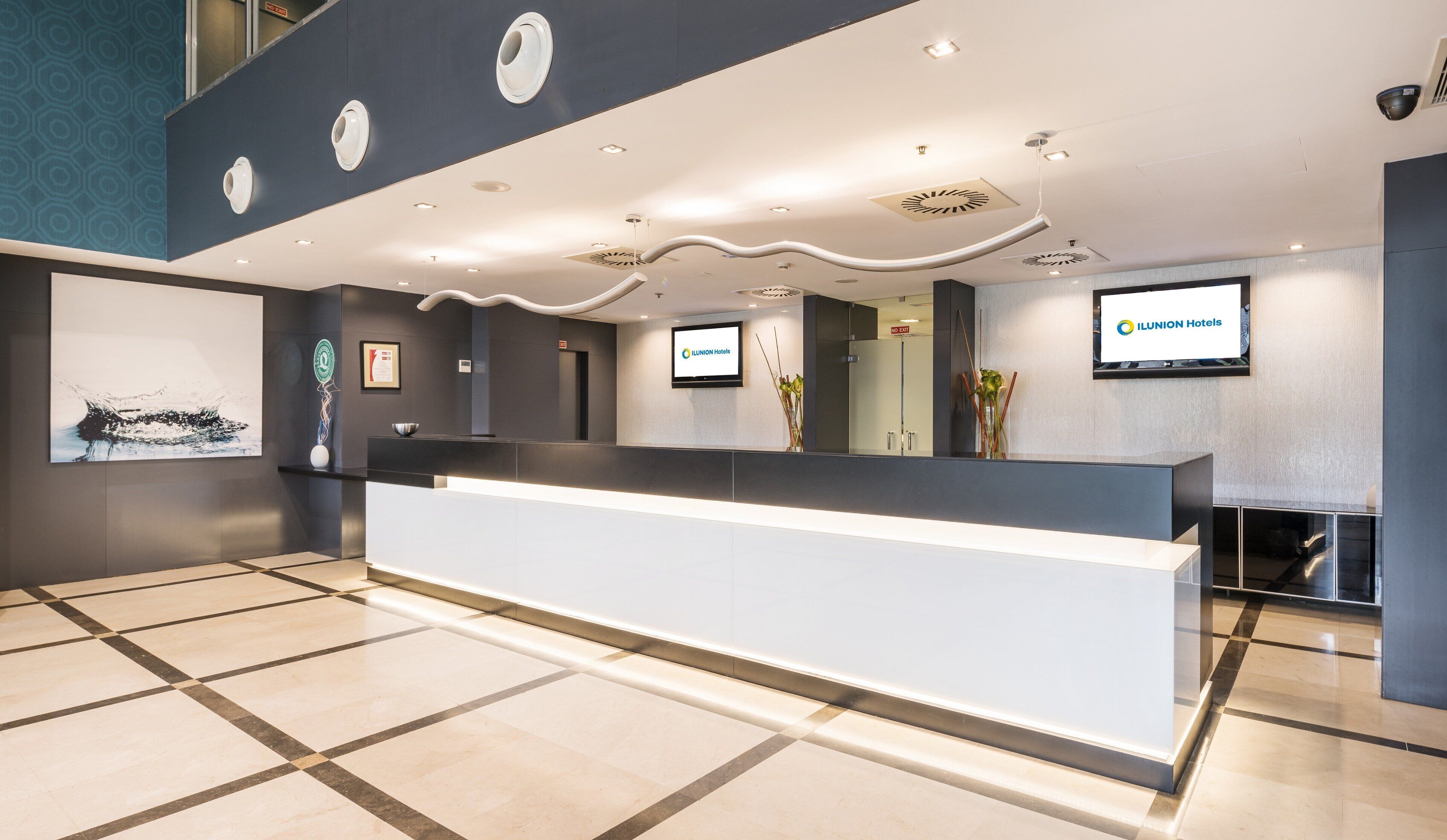 A modern hotel reception area with a large reception desk, a mounted television displaying the hotel name, and a framed certificate on the wall.