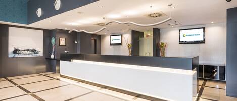 A modern hotel reception area with a large reception desk, a mounted television displaying the hotel name, and a framed certificate on the wall.
