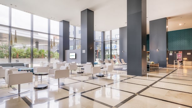 Un hall espacioso con diseño moderno, que incluye un área de descanso con sillas y sofás blancos, una pantalla digital grande y vistas al exterior.