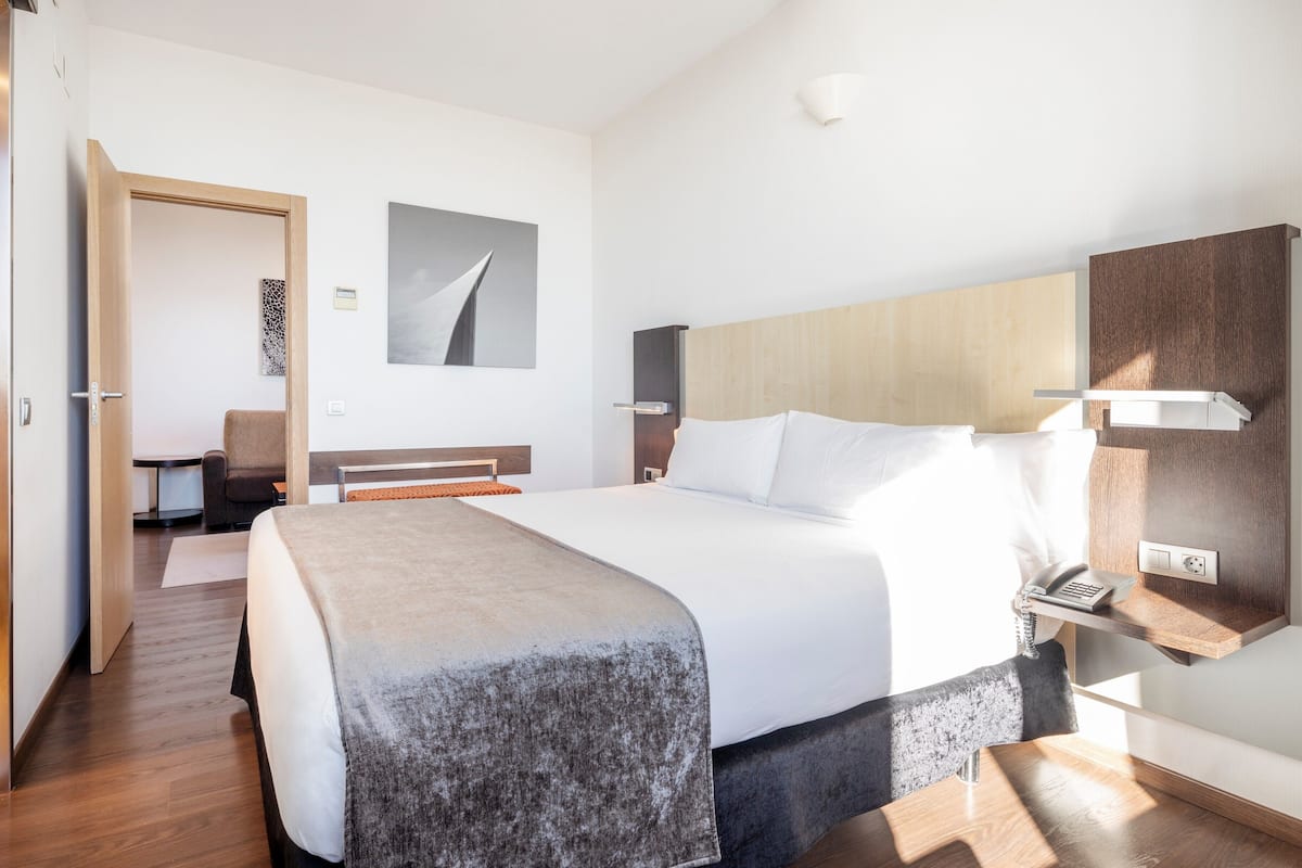 Une chambre d’hôtel moderne avec un grand lit, une tête de lit en bois, une table de chevet avec un téléphone et un petit bureau avec une chaise.