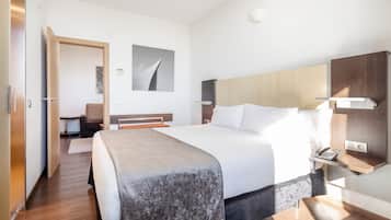 A modern hotel room with a large bed, a wooden headboard, a bedside table with a phone, and a small desk with a chair.