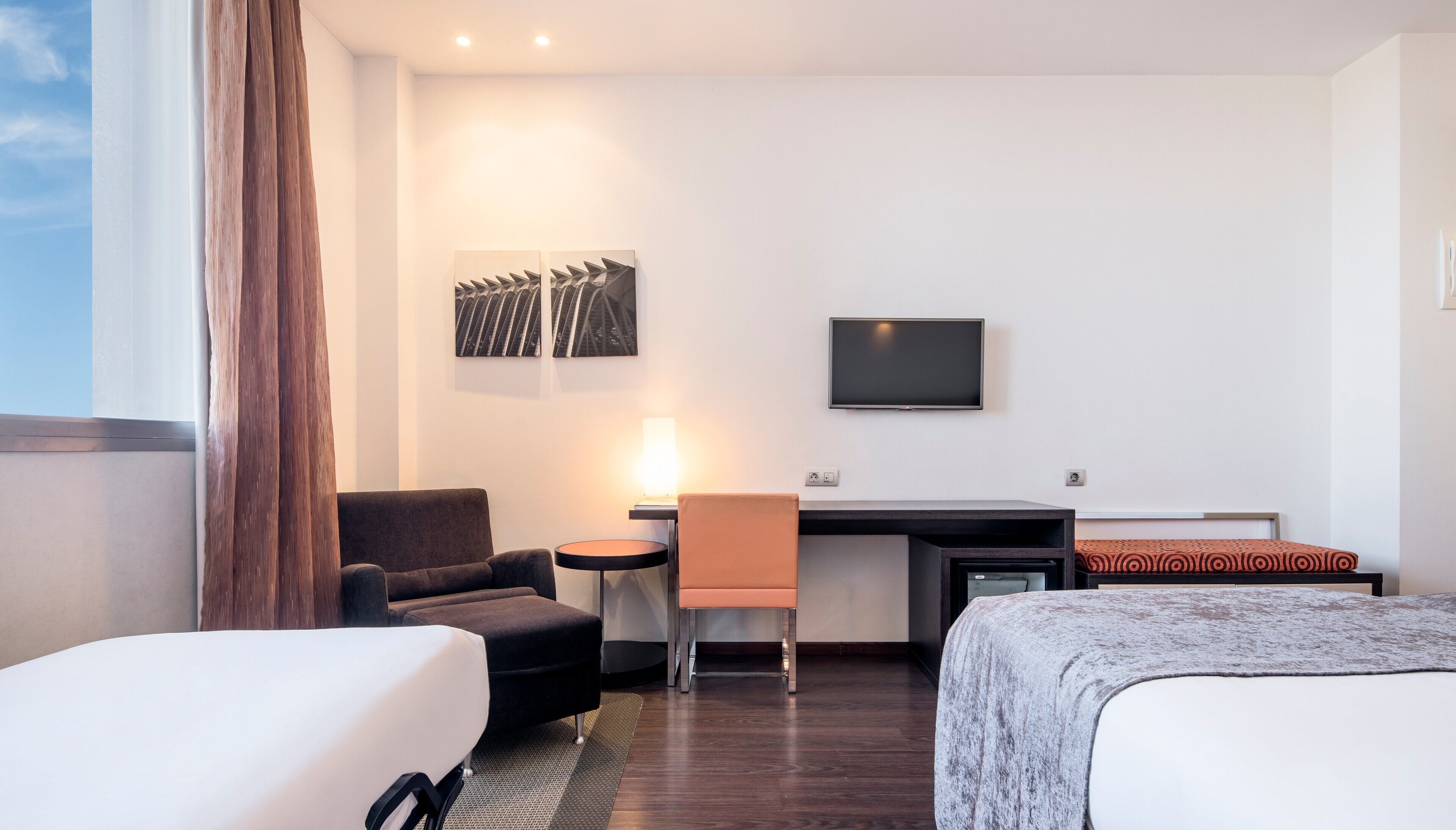 A hotel room with a bed, a desk with a chair, a TV, and artwork on the wall.