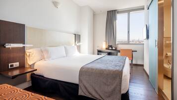 A hotel room with a bed, bedside table, lamp, and a view of the city through the window.